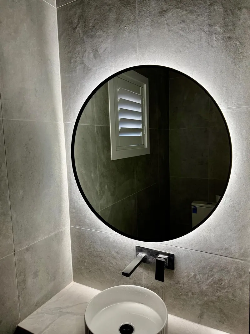 Round mirror with LED backlighting above a vessel basin in a grey tiled bathroom
