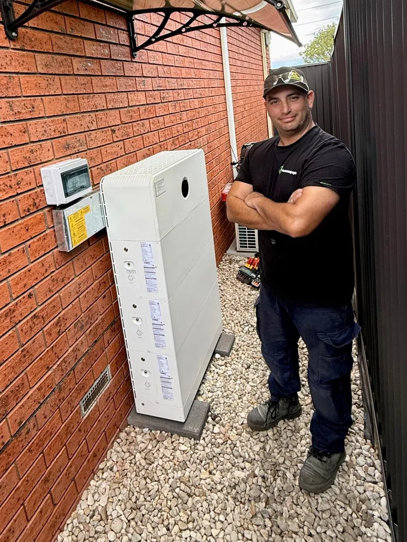 Yiannis standing arms crossed beside a stacked home battery on a brick wall