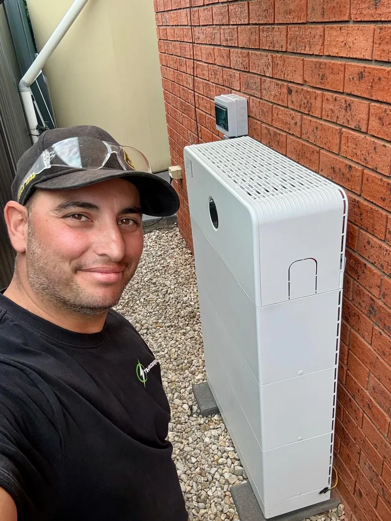 Yiannis taking a selfie beside a white battery unit installed against a brick wall