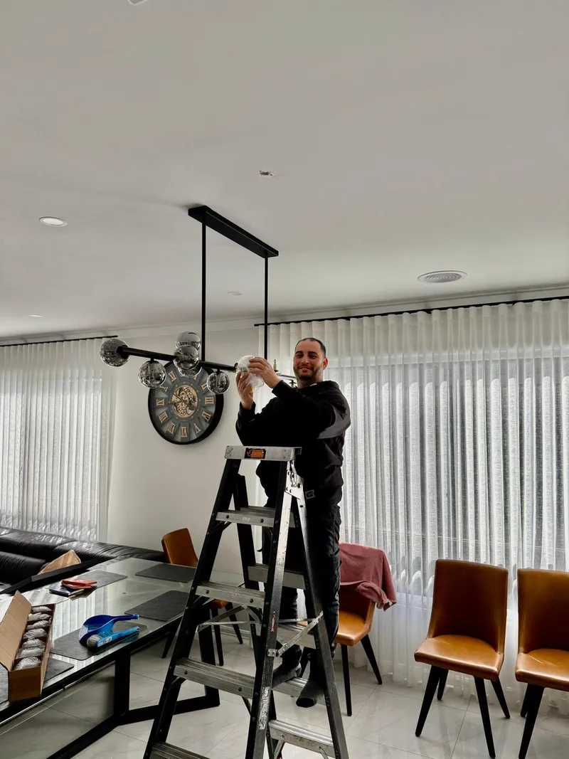 Yiannis on a stepladder fitting a multi-globe pendant light above a dining table