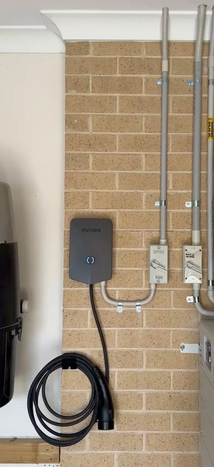 Wall-mounted EV charger with its cable coiled on a light brick wall, with conduit and isolators alongside