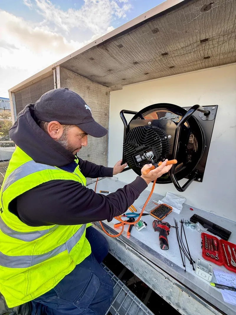 Yiannis in a hi-vis vest wiring a fan motor on a refrigerated unit, with tools laid out
