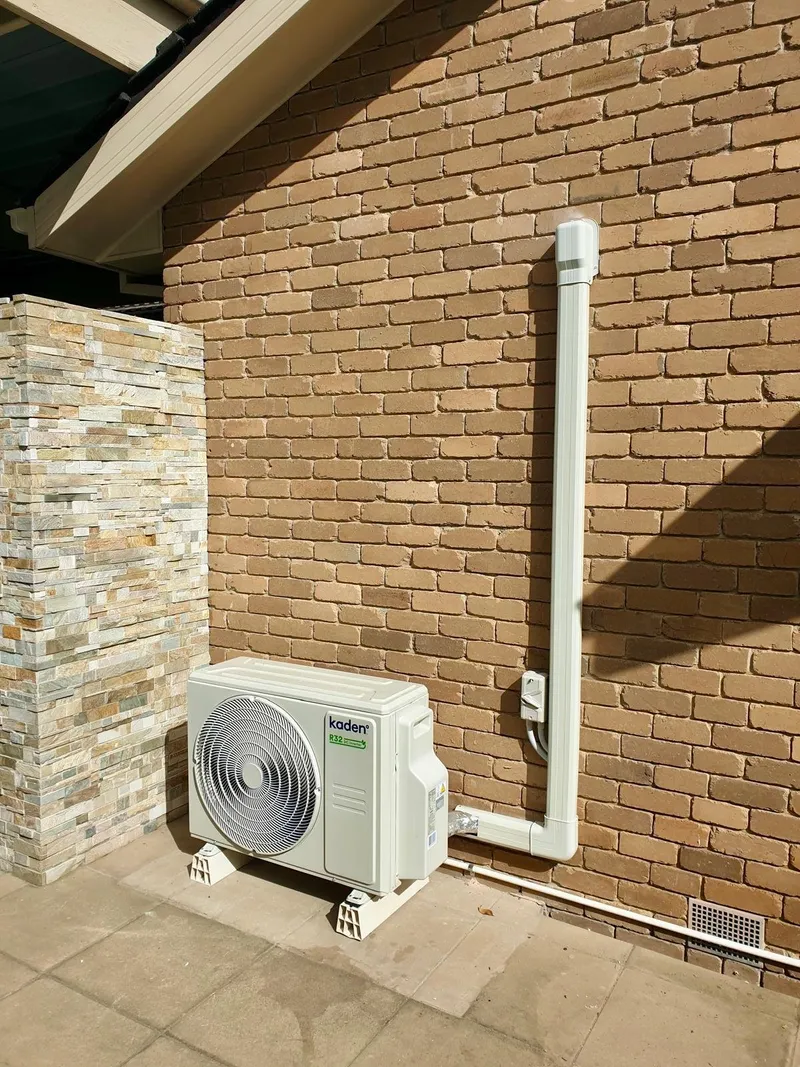Kaden outdoor unit in a paved courtyard, with a pipe cover running up the brick wall