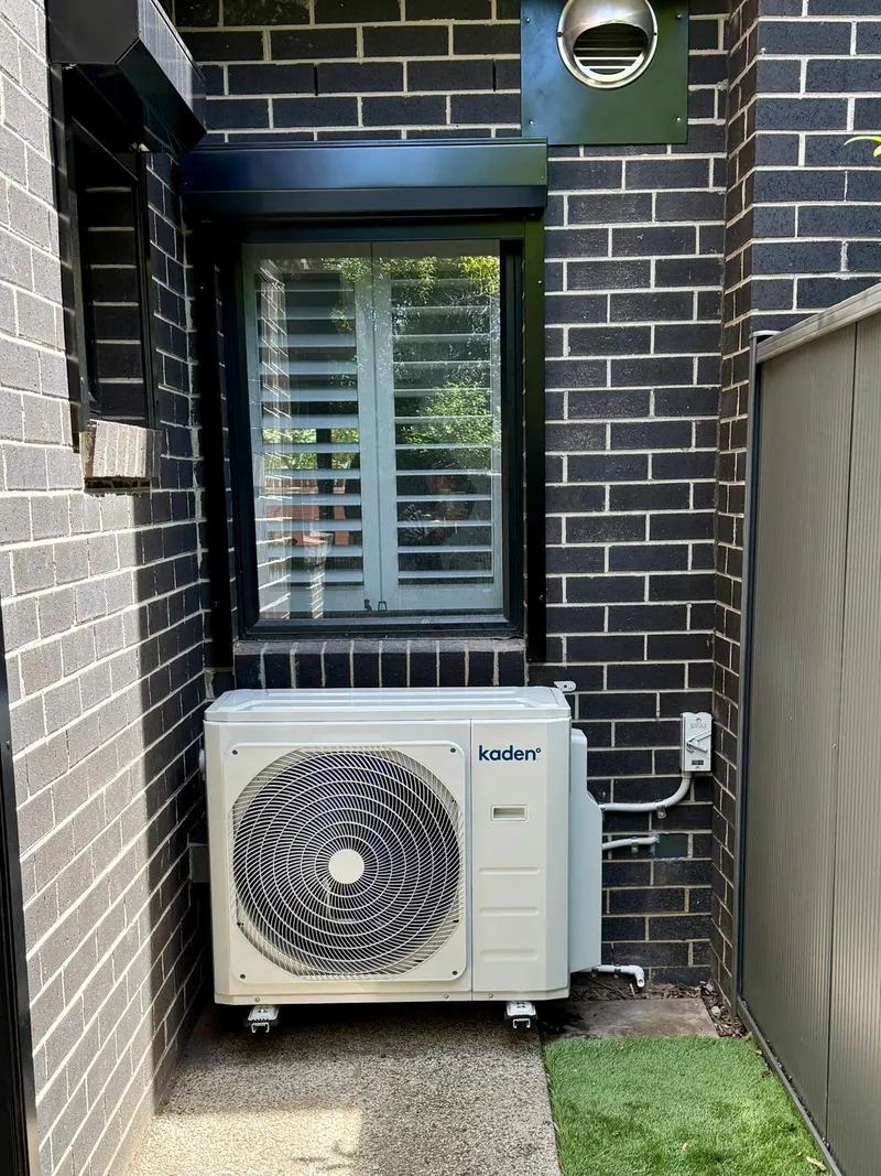 Kaden outdoor unit on a concrete path below the window of a dark brick home