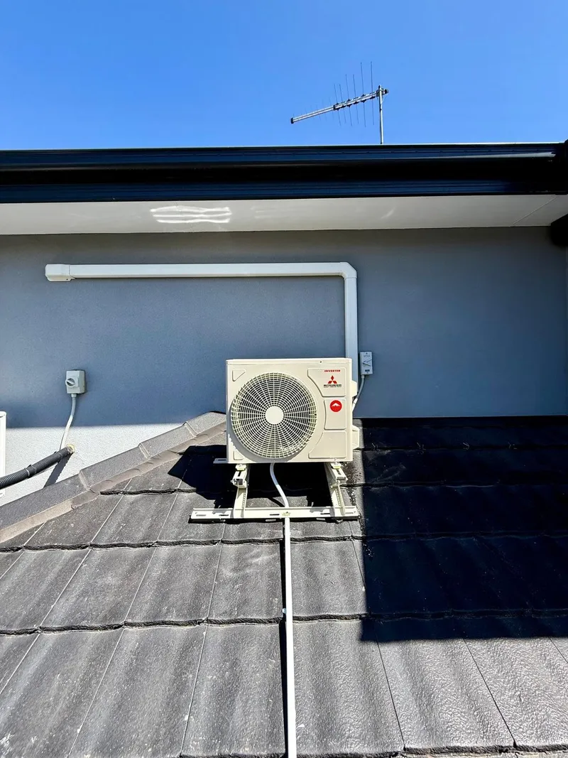 Mitsubishi outdoor unit on a roof-mount frame on a grey tiled roof, with covered pipework