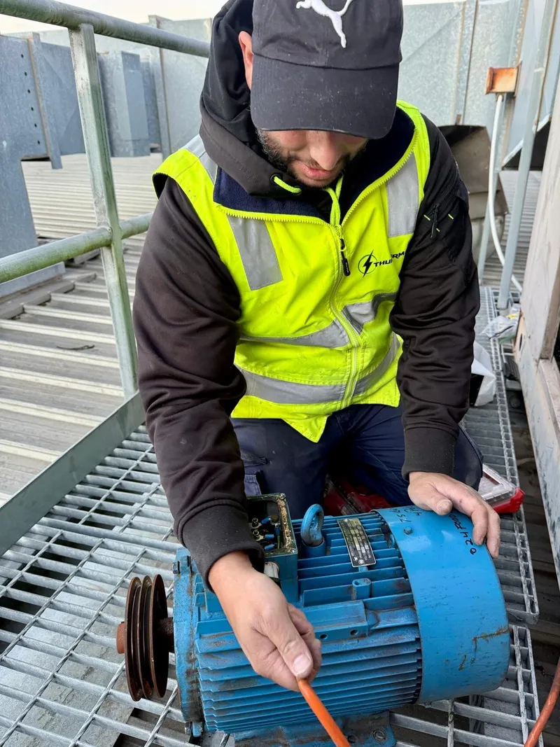 Yiannis in a hi-vis vest working on a blue electric motor on a rooftop plant platform