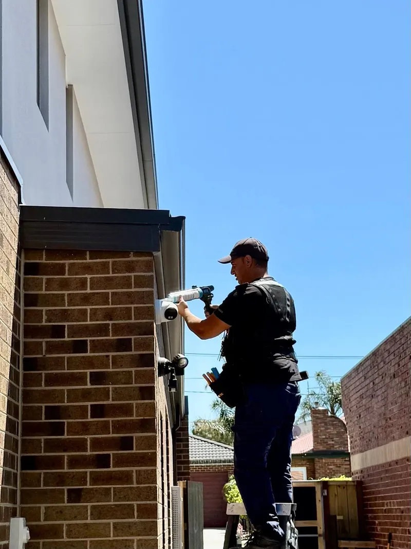 Yiannis on a ladder fitting a security camera to the brick wall of a home
