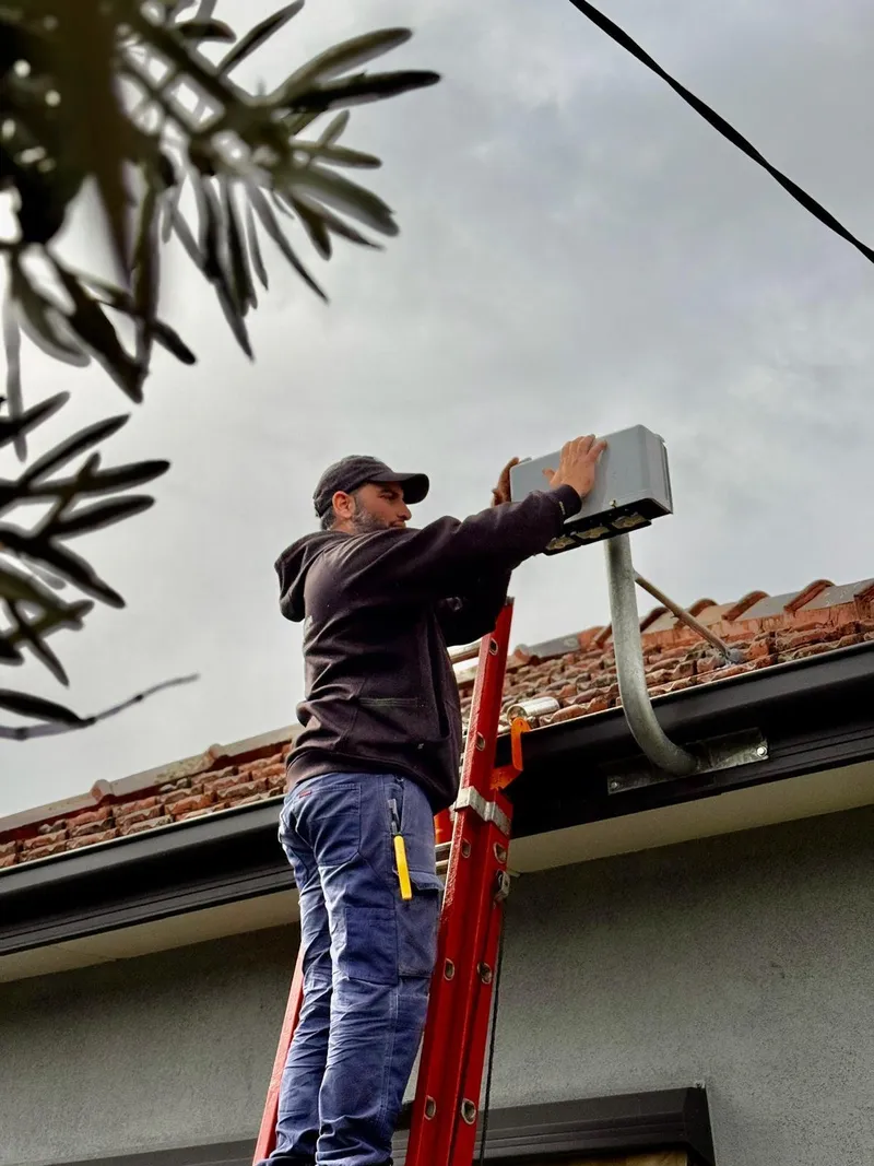 Yiannis on a ladder at the gutter line, working on a grey connection box on a galvanised mast