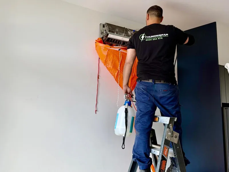 Yiannis on a ladder cleaning a wall-mounted split system, with a drain bag fitted underneath