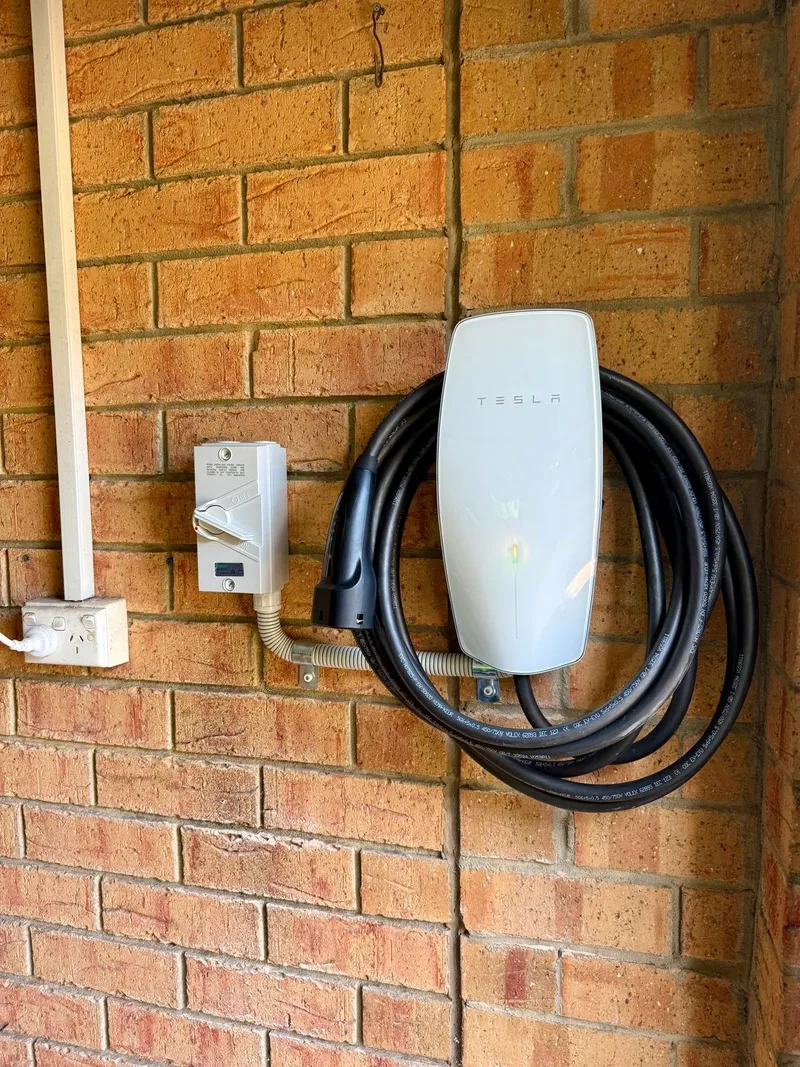 Tesla wall connector EV charger with its cable coiled, on a brick wall beside an isolator switch