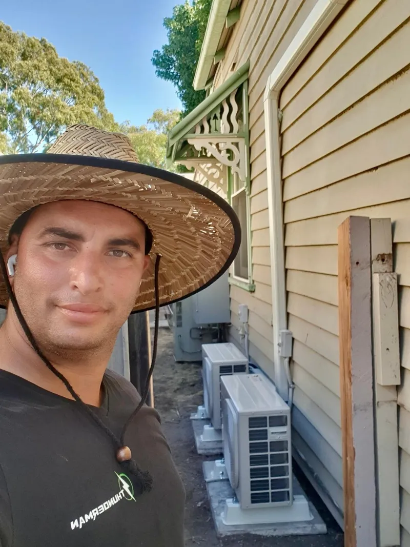Yiannis in a wide-brim hat taking a selfie beside two outdoor units