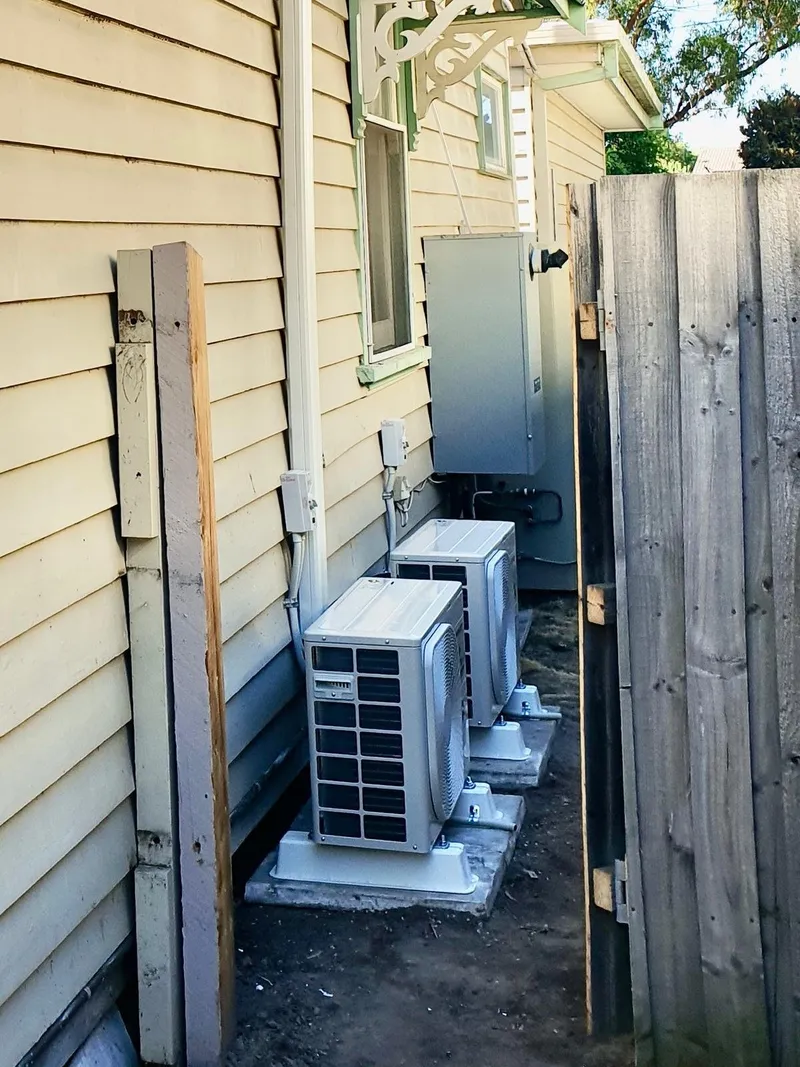 Two outdoor units on concrete pads in a narrow side passage beside a weatherboard home