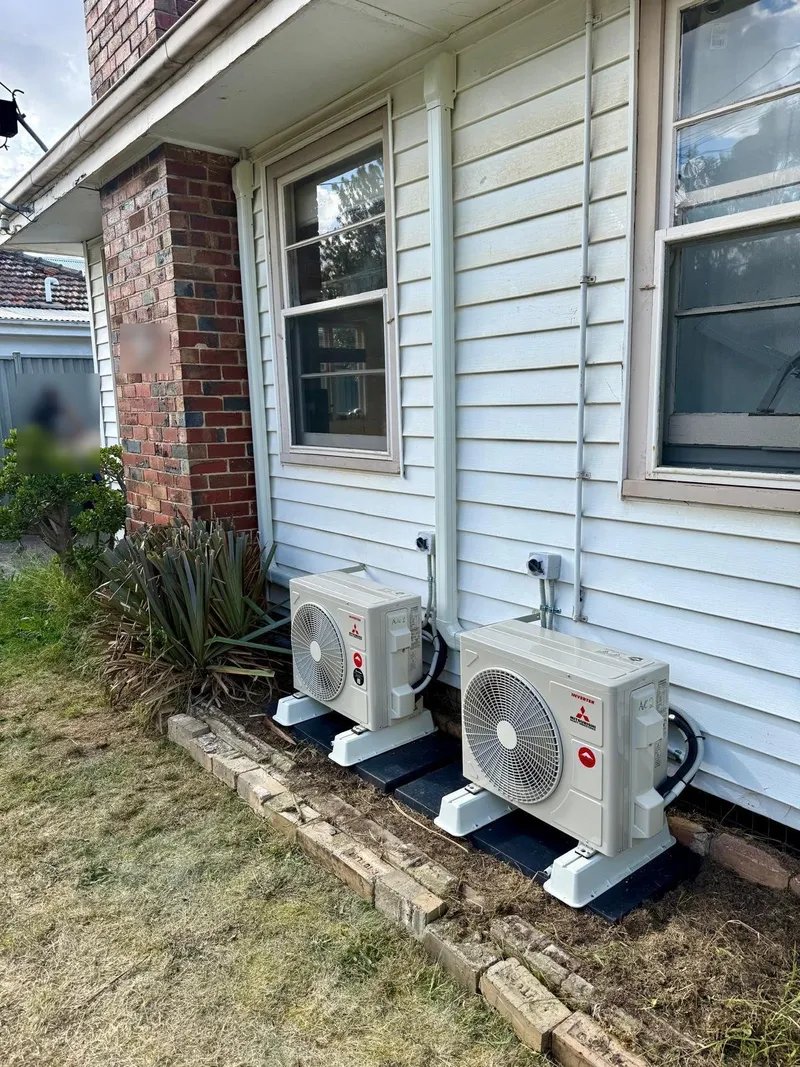 Two Mitsubishi outdoor units on stands along the side of a weatherboard home