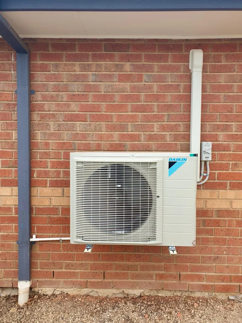 Daikin outdoor unit on wall brackets on a red brick wall, with a covered pipe run