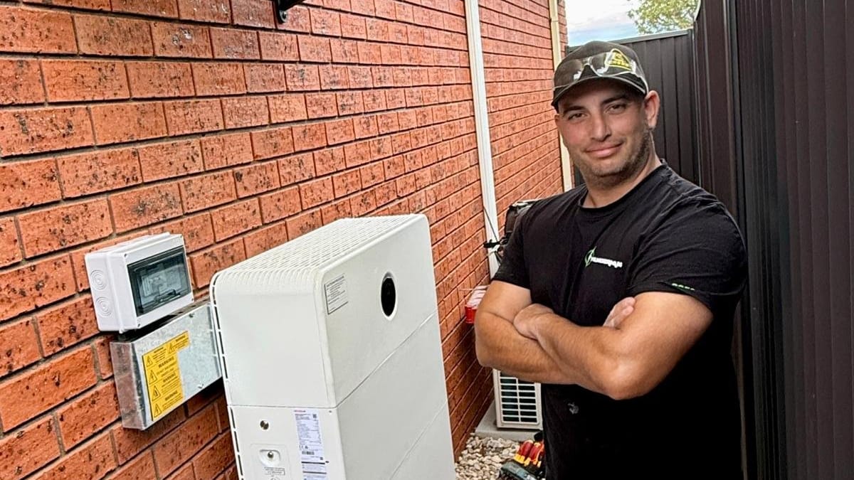 Yiannis standing arms crossed beside a stacked home battery on a brick wall