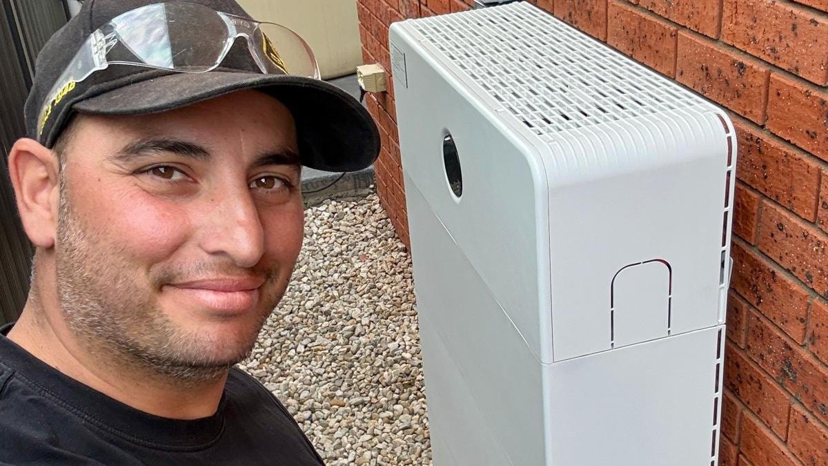 Yiannis beside a white battery unit installed against a brick wall