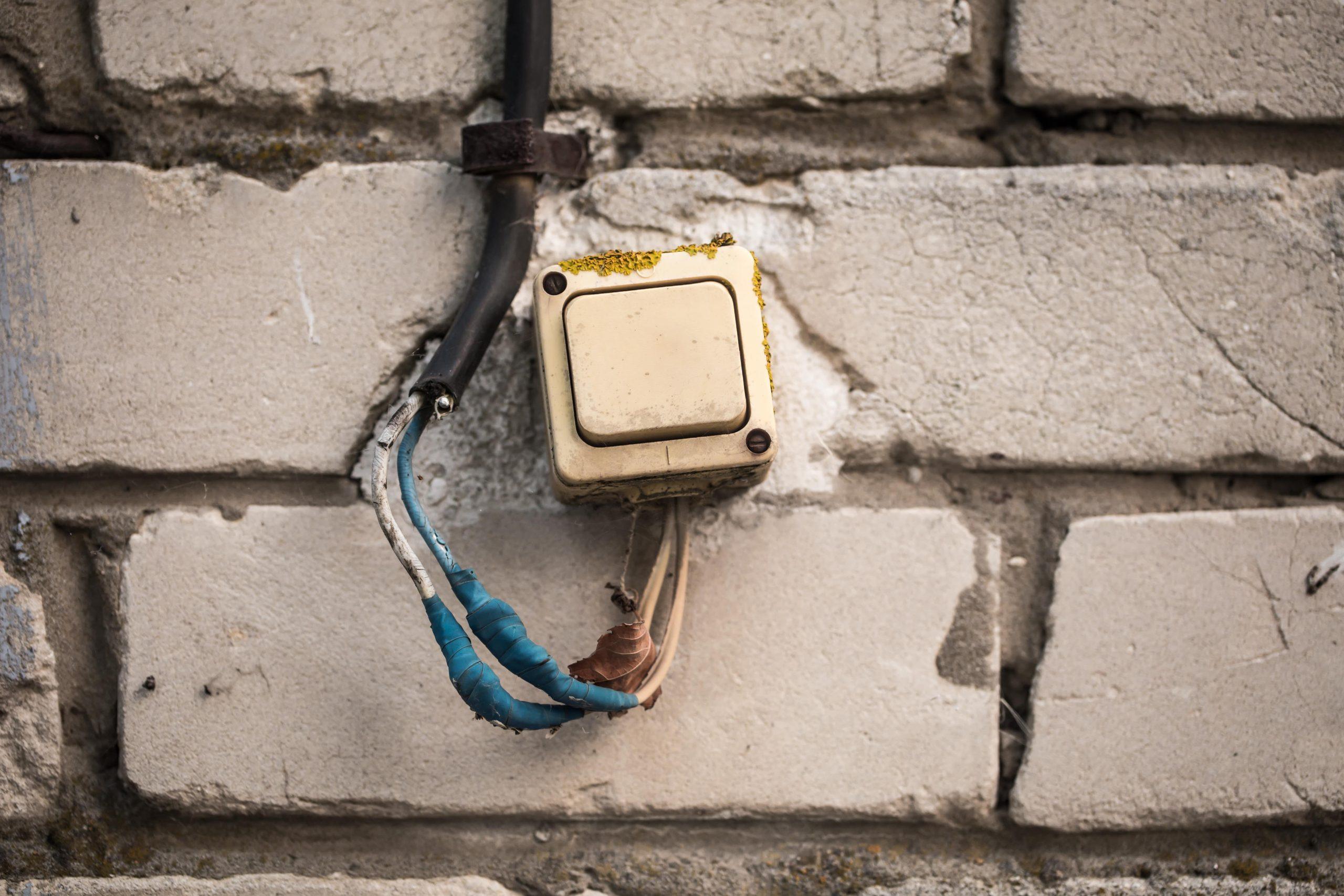 Old and damaged electrical switch box mounted on a brick wall with exposed and frayed wires covered in blue tape, highlighting potential electrical hazards.