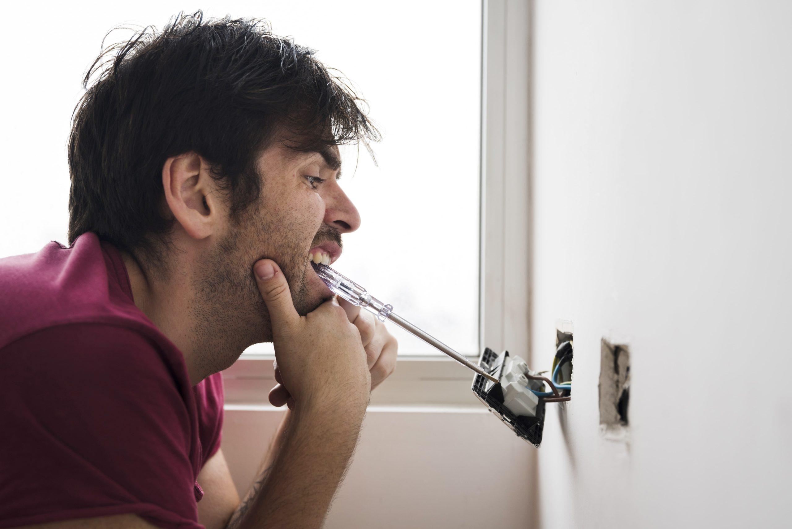 Man holding a screwdriver with his mouth while working on an exposed electrical outlet, illustrating unsafe electrical repair practices