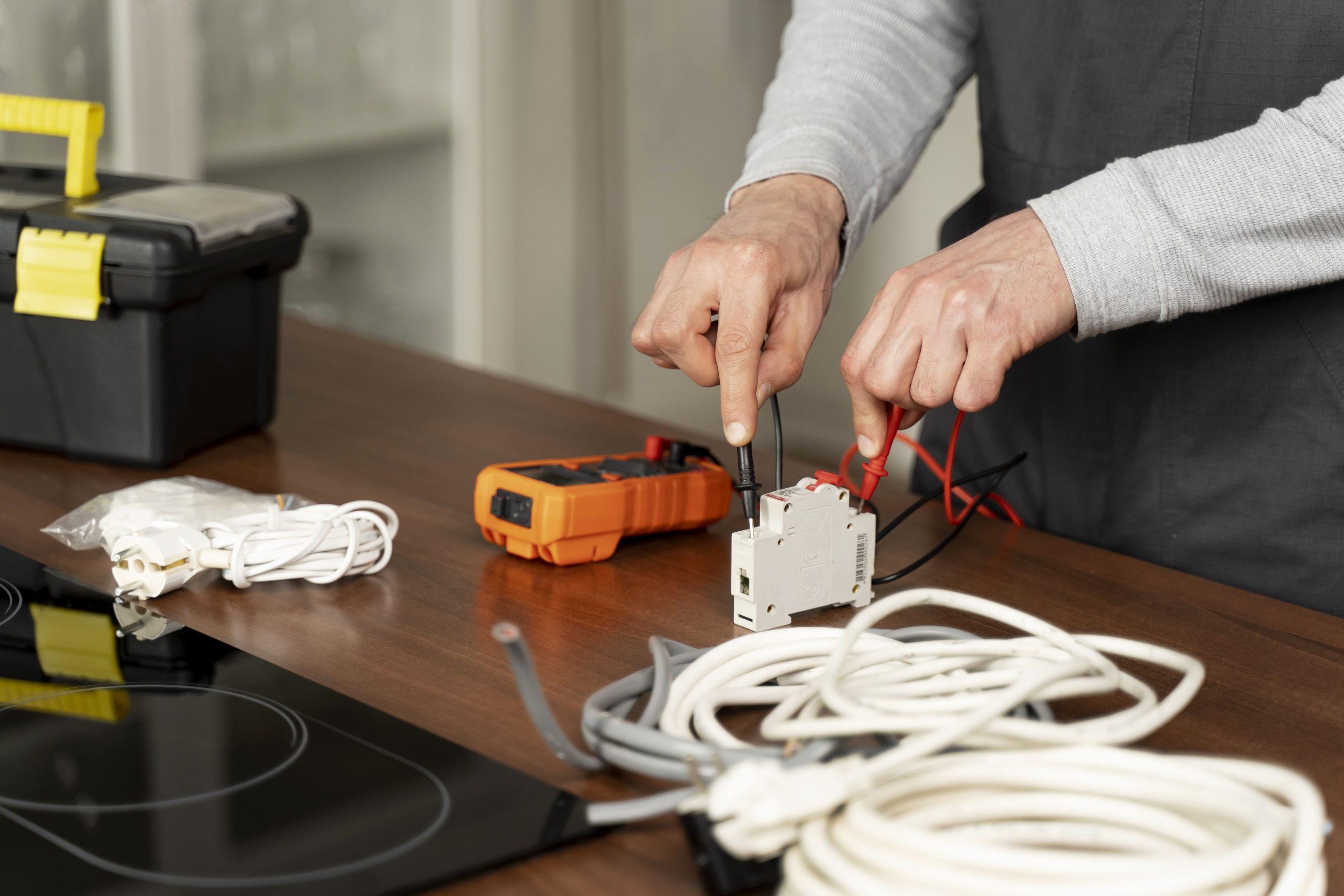 Hands testing an electrical component with a multimeter on a wooden table surrounded by wires, cables, and a toolkit, illustrating electrical maintenance work.